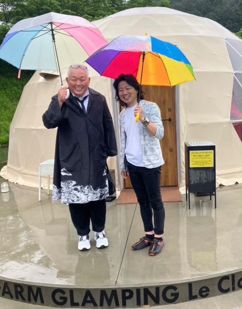 雨の日、グランピングドームの前で傘をさして並ぶ江原啓之さん(左)。足元に FARM GLAMPING Le Clos の文字が見える。