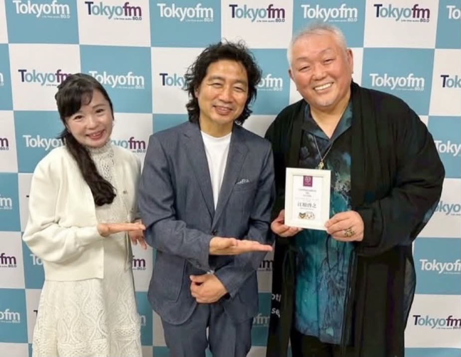 TOKYO FM のロゴが並ぶボードの前で、ユニバーサルレストラン「ル・クロ」アンバサダーの認定証を手にする江原啓之さん(右)
