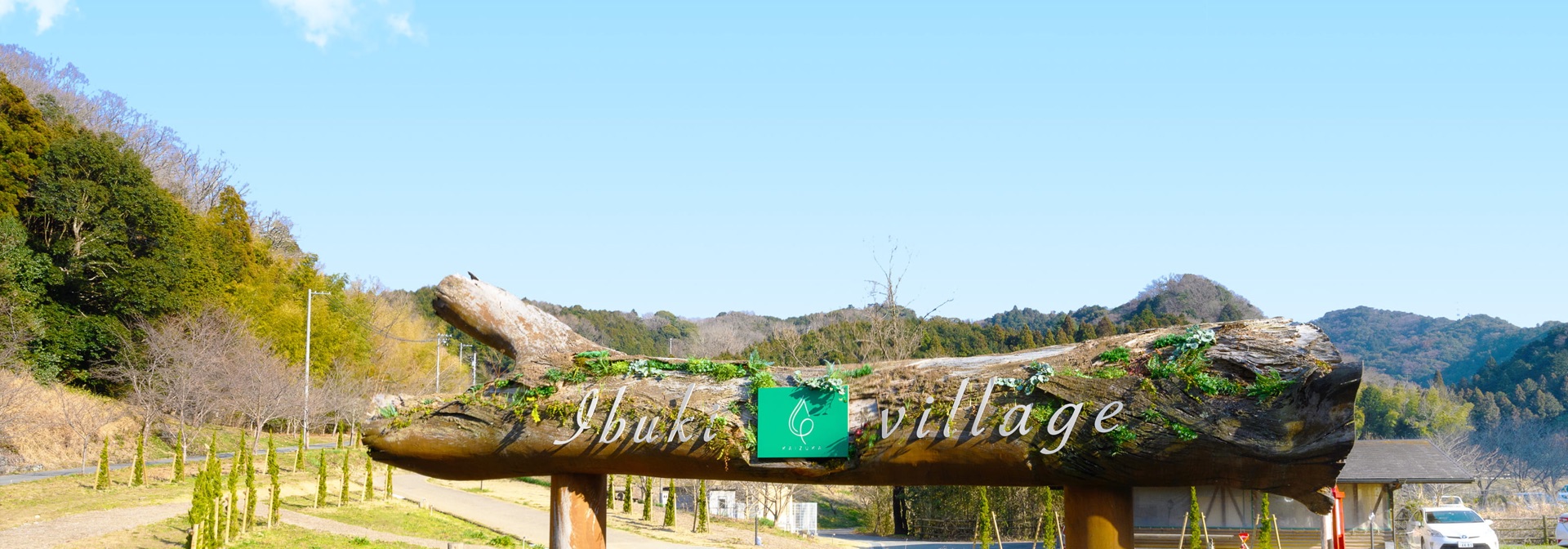 かいづか いぶきヴィレッジのエントランス。丸太に Ibuki village と彫られた看板と、奥に広がる里山